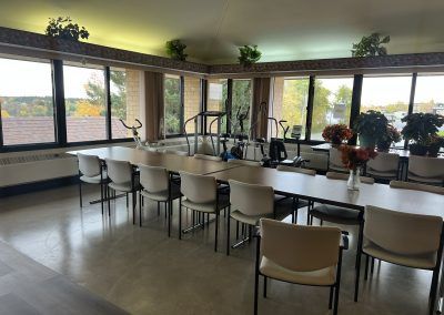 Common area long table with chairs and exercise equipment in corner of main room with lots of windows in apartment building.